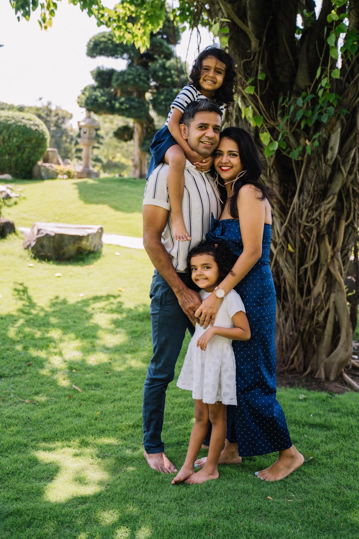 A family of four smiling together outdoors under a tree